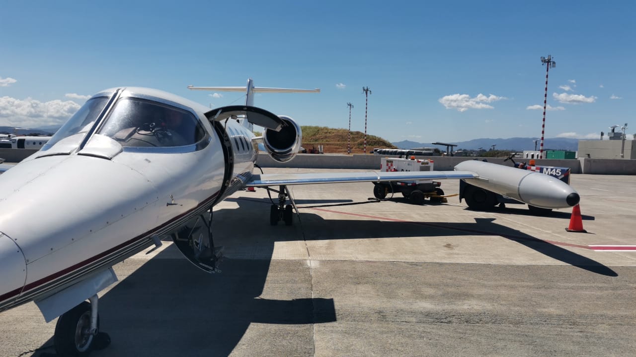 Close-up view of a white private jet on an airport ramp with its door open and engine visible, a second jet in the background, and ground equipment nearby under a blue sky.